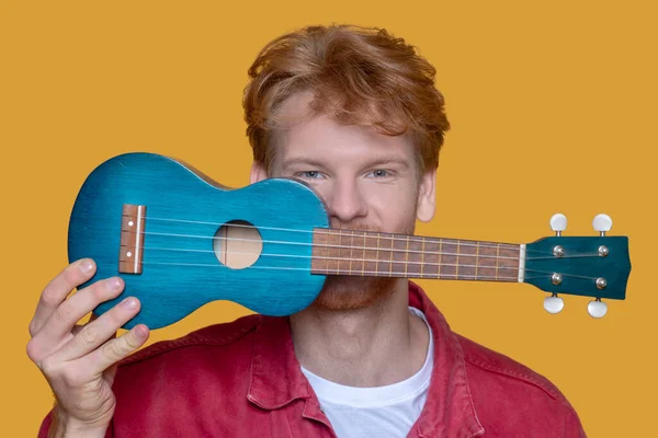 Jovem barbudo homem de boa aparência em casaco vermelho com ukulele se divertindo — Fotografia de Stock