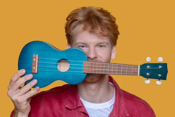 Young bearded good-looking man in red jacket with ukulele having fun — Stock Photo, Image