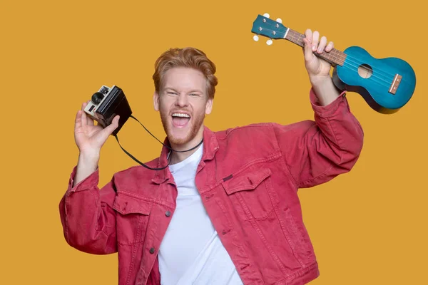 Young bearded good-looking man in red jacket with ukulele and camera — Stock Photo, Image