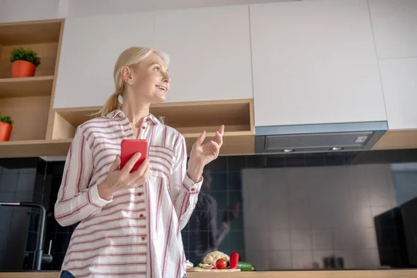 Mujer de pie en la cocina con un teléfono inteligente en la mano y mirando reflexivo — Foto de Stock
