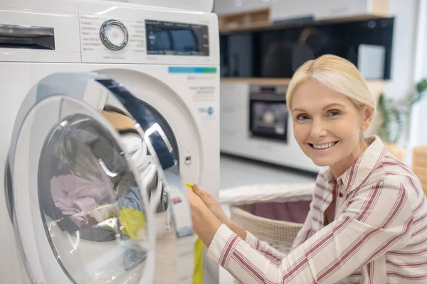 Mujer rubia poniendo ropa en la lavadora y sonriendo —  Fotos de Stock