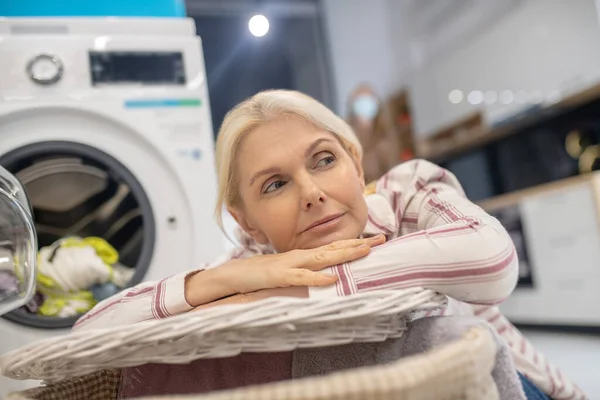 Mujer rubia sentada cerca de la lavadora y buscando reflexivo —  Fotos de Stock