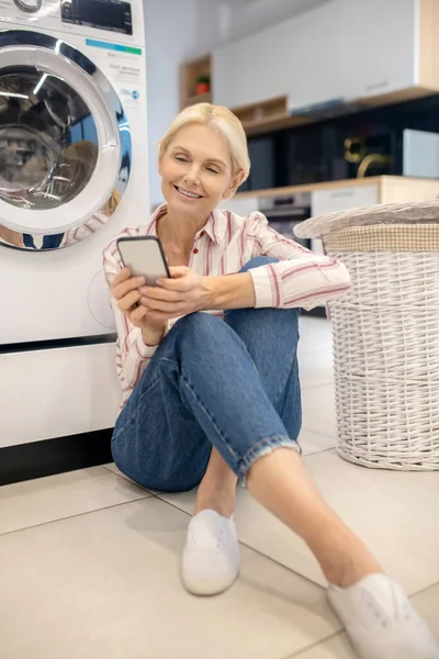 Rubia ama de casa con camisa a rayas sentada cerca de la lavadora y leyendo algo en línea —  Fotos de Stock