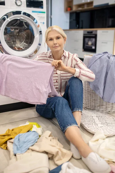 Amante rubia con camisa a rayas sentada cerca de la lavadora y sosteniendo la ropa —  Fotos de Stock