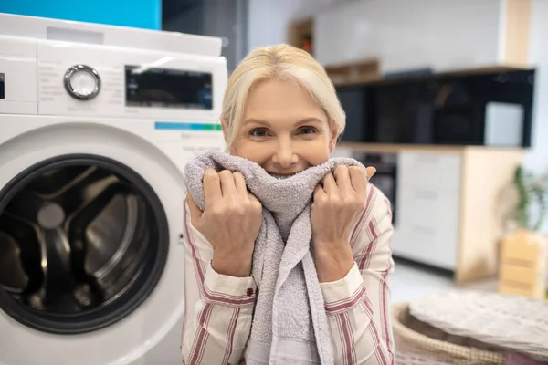 Blond huisvrouw in gestreept shirt zit naast de wasmachine en ruikt schone kleren — Stockfoto