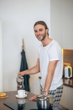 Ev elbiseli genç adam fincana kahve dolduruyor ve gülümsüyor.