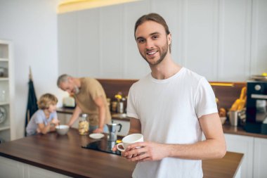 Beyaz tişörtlü genç adam mutfakta duruyor ve gülümsüyor.