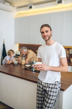 Beyaz tişörtlü genç adam mutfakta duruyor ve güzelce gülümsüyor.