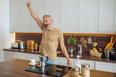 Bej tişörtlü olgun bir adam mutfakta duruyor ve esniyor.