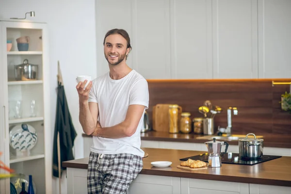 Hombre joven en ropa de casa de pie en la cocina con una taza de café en la mano — Foto de Stock