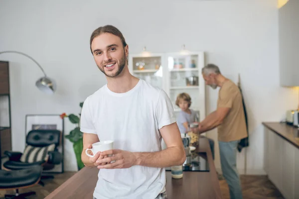 Ung man i vit tröja som står i köket och ler — Stockfoto