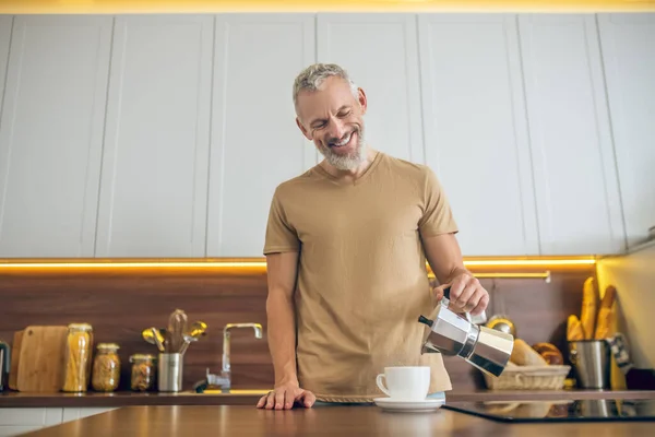 Volwassen man in beige tshirt gieten koffie naar de beker — Stockfoto