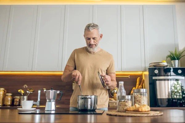 Hombre maduro en camiseta beige de pie en la cocina y haciendo el desayuno —  Fotos de Stock