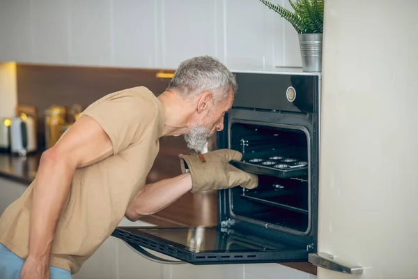 Hombre maduro en camiseta beige sacando algo del horno — Foto de Stock