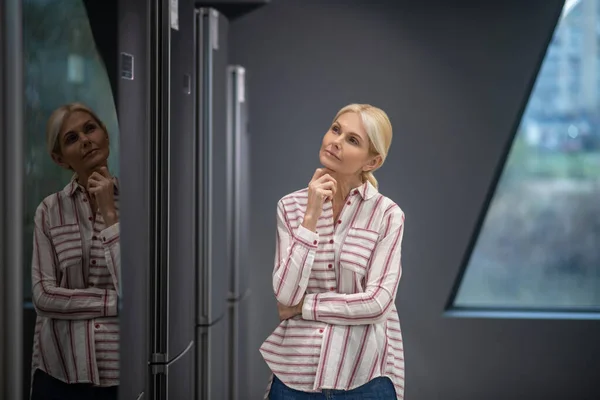 Женщина в полосатой рубашке выбирает холодильник в выставочном зале и выглядит заботливо — стоковое фото