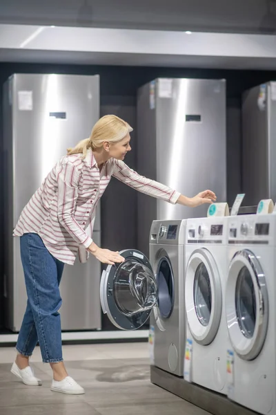 Donna in camicia a righe scegliendo una lavatrice in uno showroom e cercando interessati — Foto Stock