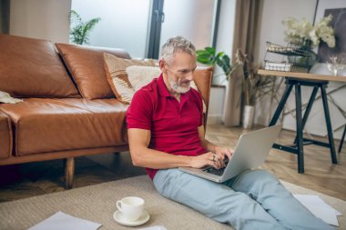 Gri saçlı adam yerde oturuyor ve dizüstü bilgisayarda yazıyor.