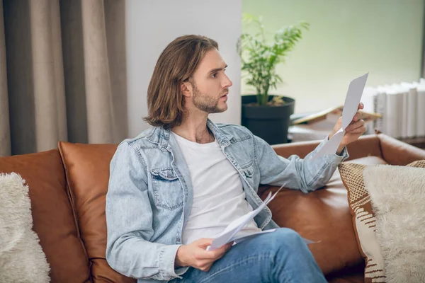 Young man in jeans shirt holding papers and looking thoughtful — Stock Photo, Image