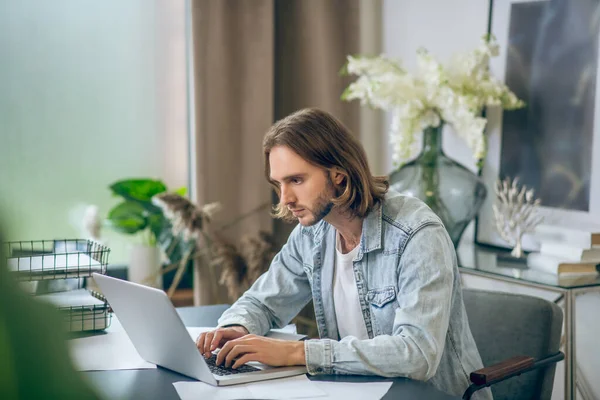 Hosszú hajú férfi farmeringben, laptopon dolgozik és koncentráltnak tűnik. — Stock Fotó