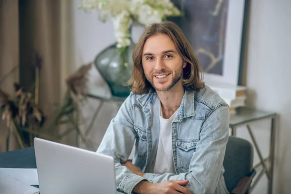 Långhårig man i jeans skjorta arbetar på laptop och ser glad ut — Stockfoto