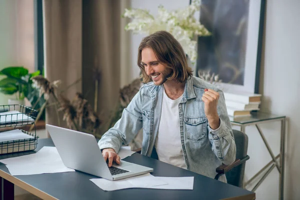 Lange haar man in jeans shirt werken op laptop en op zoek gelukkig — Stockfoto