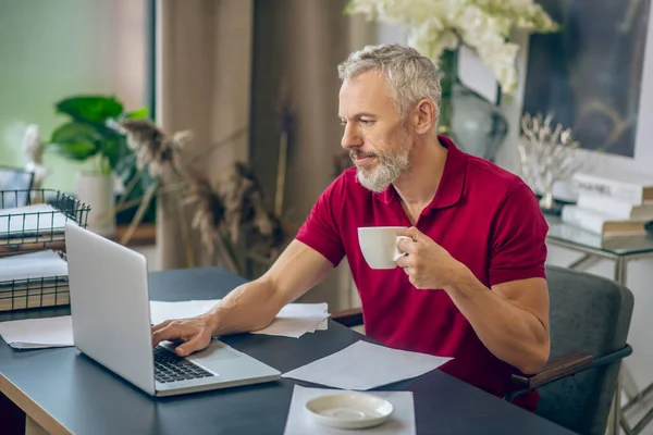 회색 머리 남자가 노트북 앞에 앉아서 커피를 마시고 있습니다. — 스톡 사진