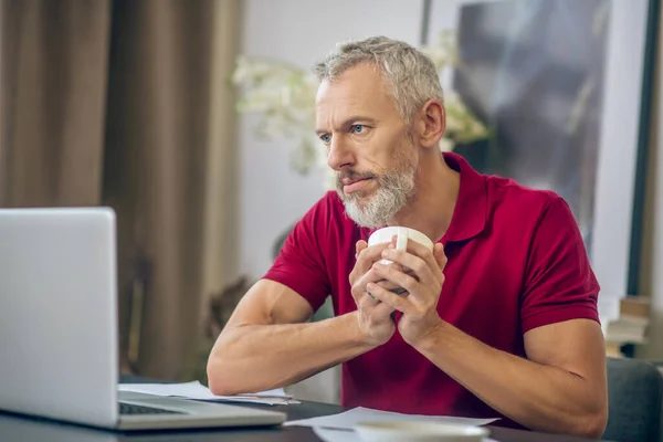 Gri saçlı adam dizüstü bilgisayarda oturuyor ve kahve içiyor. — Stok fotoğraf