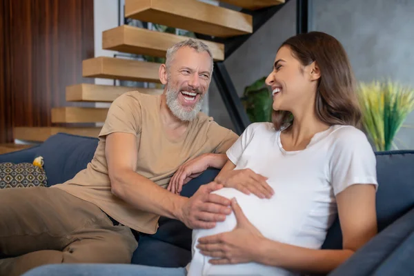Homem maduro feliz tocando abdômen de sua jovem esposa grávida e olhando feliz — Fotografia de Stock