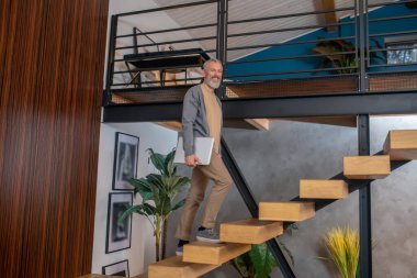 Gri saçlı, olgun, kendine güvenen bir adam elinde dizüstü bilgisayarla yukarı çıkıyor.