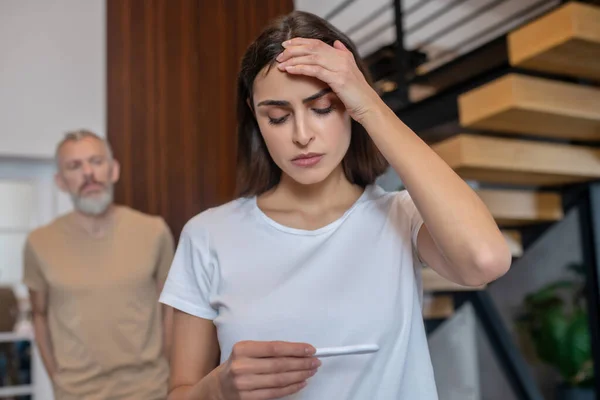 Ung mörkhårig kvinna tittar stressad medan du tittar på graviditetstestet och prata med sin man — Stockfoto