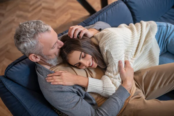 Homem de cabelos grisalhos sentado no sofá e abraçando sua esposa com ternura — Fotografia de Stock