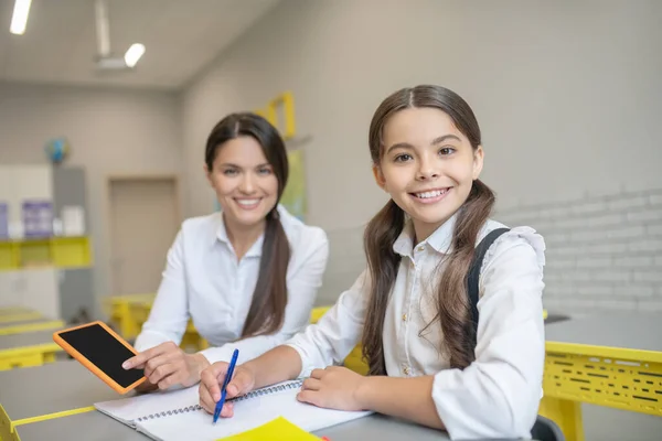 Smiling teacher and schoolgirl sitting at desk — Stock Photo, Image