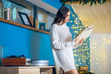 Siyah saçlı, beyaz elbiseli bir kadın elinde bir plak tutuyor.