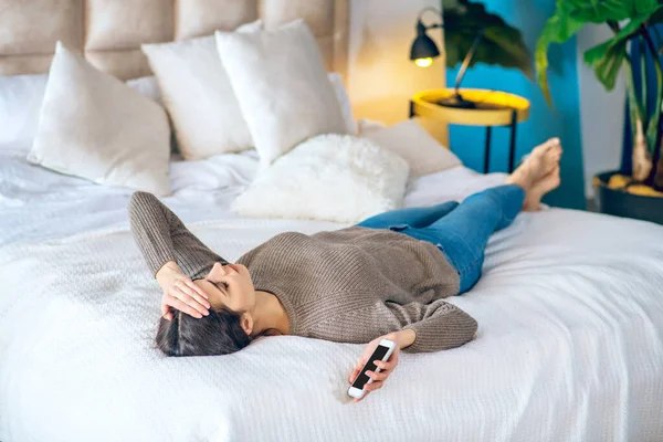 Темноволоса молода жінка лежить на ліжку з телефоном в руці і виглядає нещасною — стокове фото