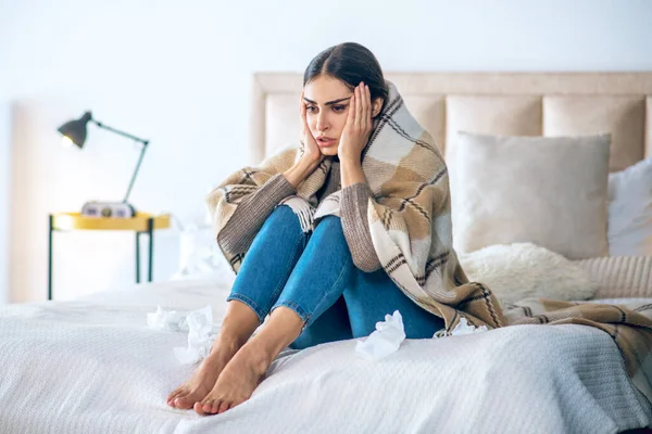 Wanita muda berambut gelap duduk di tempat tidur dan memegang kepalanya — Stok Foto