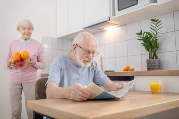 Senior grey-haired man sitting at the table in the kitchen and reading a newspaper — Stock Photo, Image