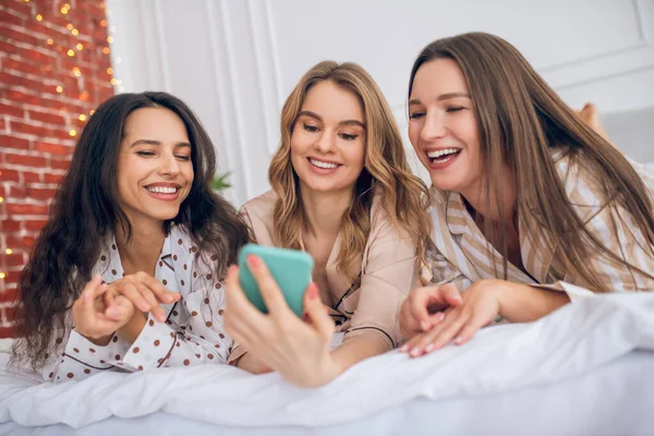 Tiga gadis berbaring di tempat tidur dan menonton sesuatu di smartphone — Stok Foto