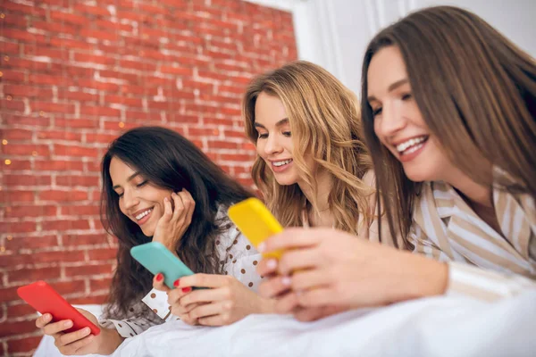 Chicas lindas con teléfonos inteligentes tumbados en la cama y viendo algo —  Fotos de Stock