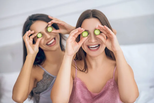 Dos chicas jóvenes lindas con rodajas de pepino en las manos que tienen funa nd mirando feliz — Foto de Stock