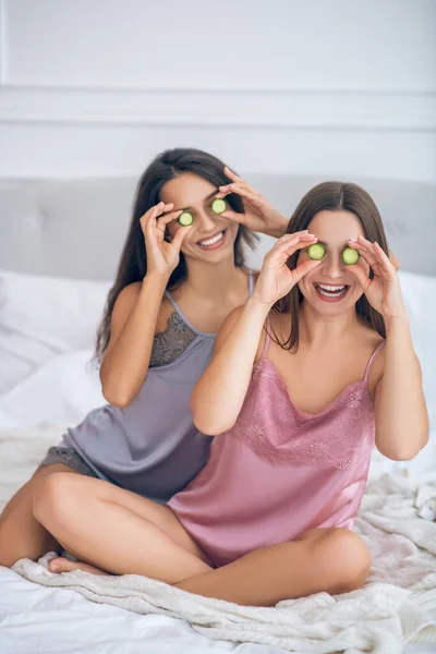 Duas meninas bonitos com fatias de pepino em mãos tendo diversão nd olhando feliz — Fotografia de Stock