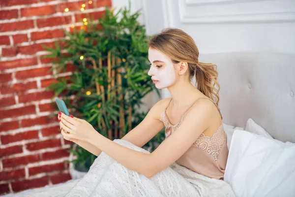 Gadis pirang dengan topeng di wajahnya duduk di tempat tidur dengan smartphone di tangan — Stok Foto