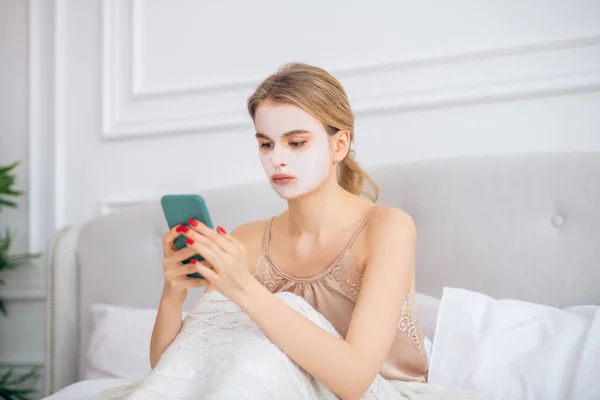 Gadis pirang dengan masker di wajahnya duduk di tempat tidur — Stok Foto