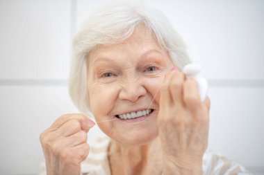 Gri saçlı kadın dişlerini temizliyor ve diş ipi kullanıyor.