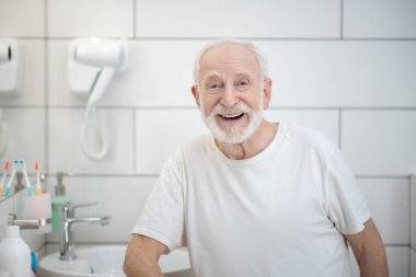 Beyaz tişörtlü, gri saçlı adam güçlü bir şekilde gülüyor.
