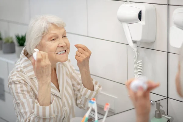 Gri saçlı kadın dişlerini temizliyor ve diş ipi kullanıyor. — Stok fotoğraf