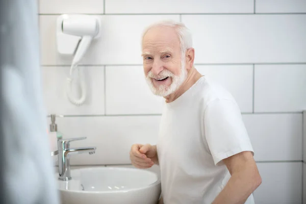 Le gråhårig man i vit tröja i badrummet — Stockfoto