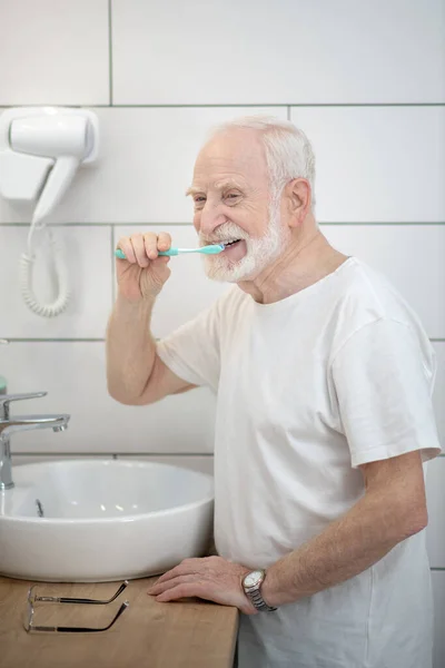 Gråhårig man i vit tröja som rengör tänderna — Stockfoto