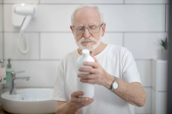 Сіроволосий чоловік в окулярах вивчає пляшку шампуню — стокове фото