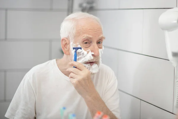 Ősz hajú férfi fehér pólóban borotválkozik a fürdőszobában, és koncentráltan néz ki. — Stock Fotó