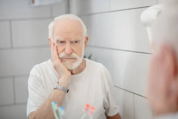 Gray-haired man in white tshirt looking in the mirror and looking worried — Stock Photo, Image